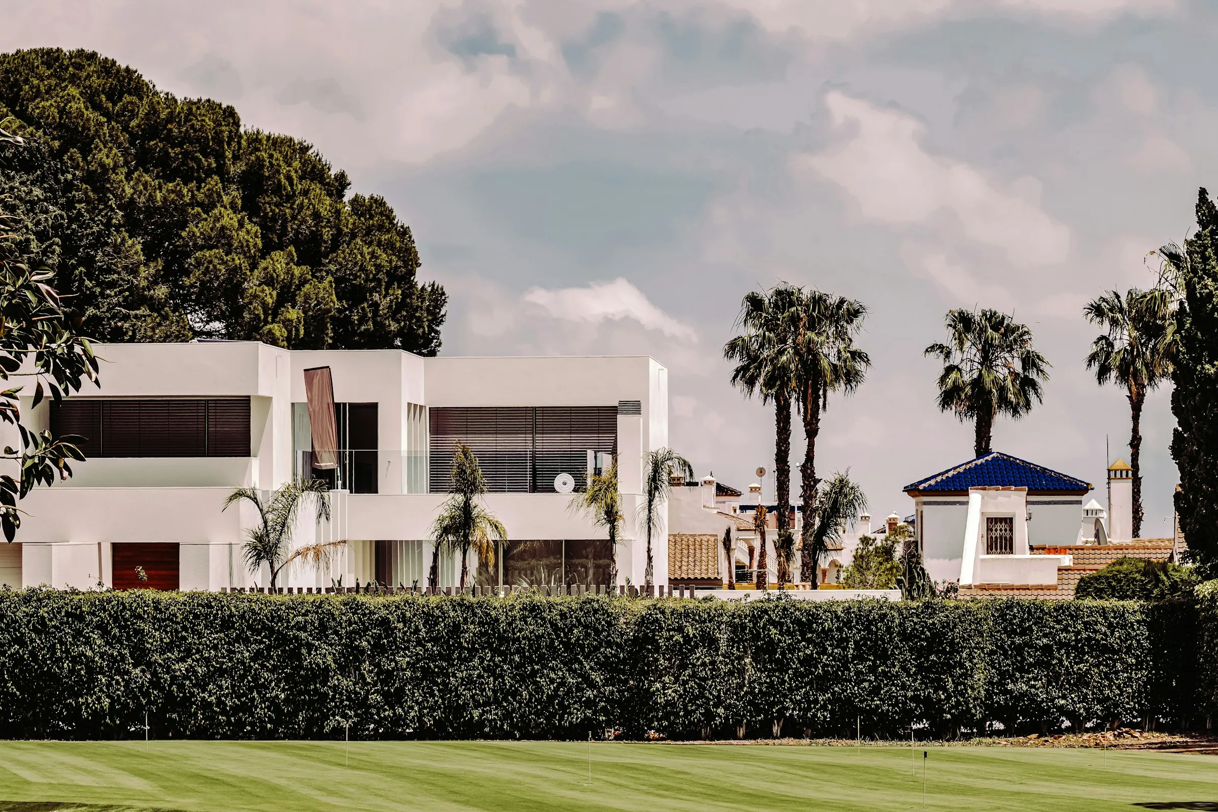 White modern villa with palm trees behind a manicured hedge